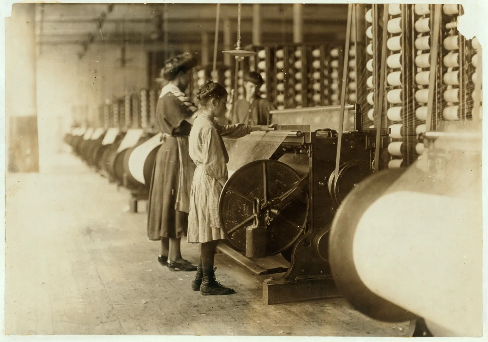 A black and white photograph of a woman, a young girl, and a tall boy working in front of a large, circular machine. There are many threads connected to the machine from a tower of spools of yarn. There are many machines of the same nature seen in rows in the background of the photograph.