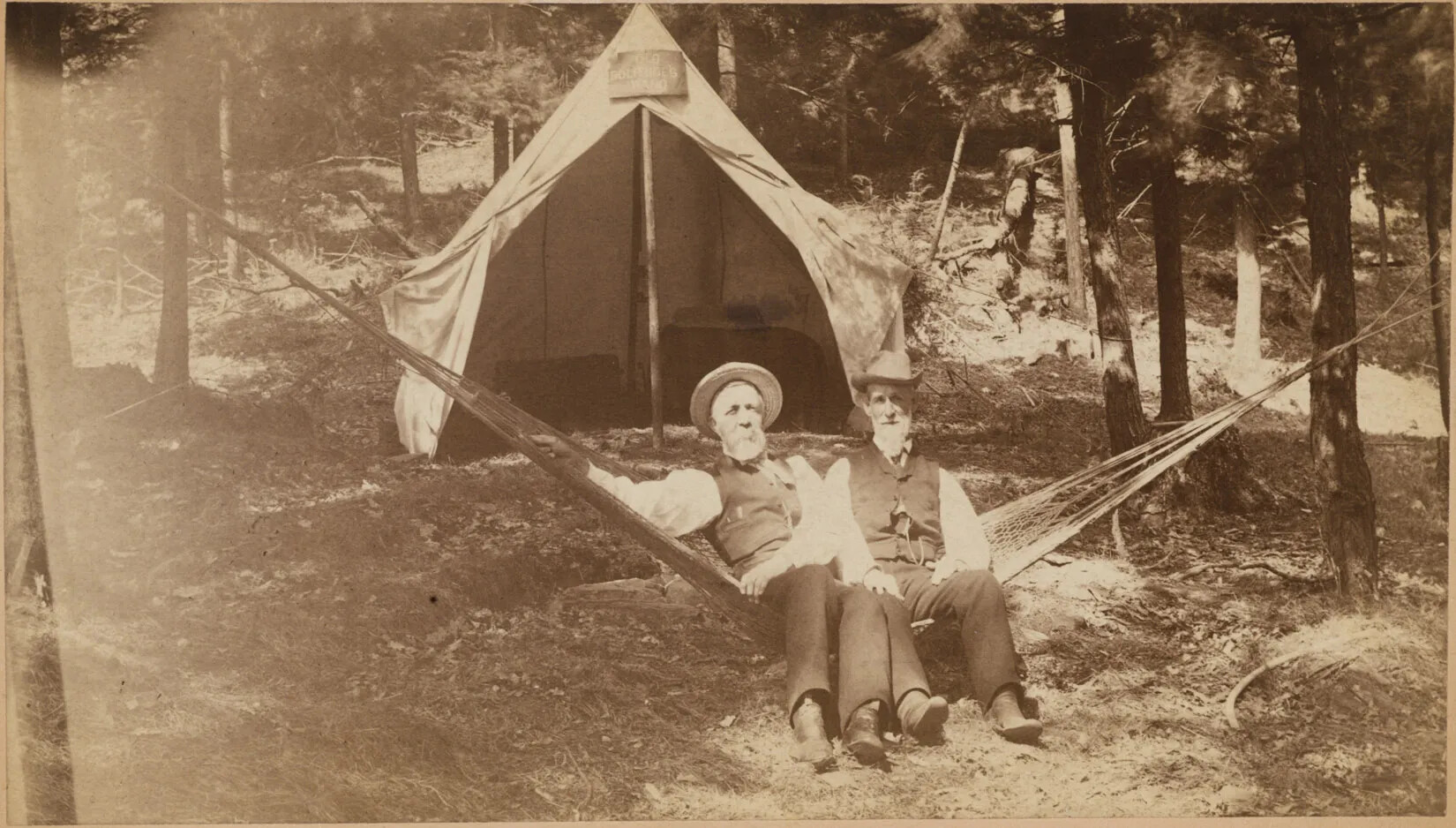 A black and white photograph of two men in a hammock in a forest. The men have white beards, and wear button-down shirts with vests, pants, and summer hats. Behind them is a canvas tent whose doors are held open by rope. Two low bunks are seen inside. 