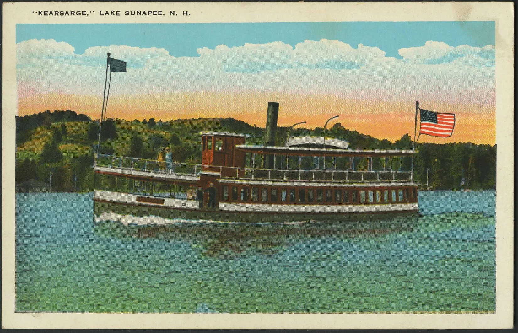 A colored postcard of a wooden steam boat on a body of water. The boat has one small flag at the front, and a large American flag on the back. In the background are trees and open grassy areas. Orange and pink clouds mix with white clouds on the blue sky. At the top-left corner are the words "'KEARSARGE,' LAKE SUNAPEE, N. H."