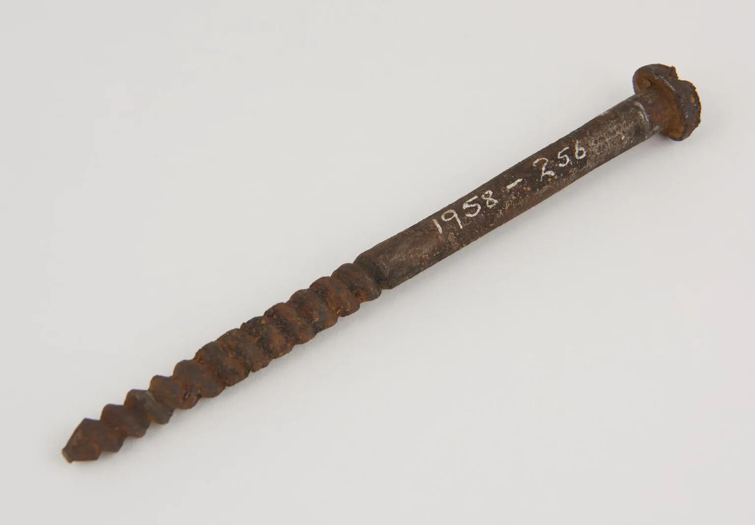 A rusty metal screw, with a circular nail head on one side and a coiled pointed end on the other. The numbers "1958 - 256" are painted on to the screw in white lettering.