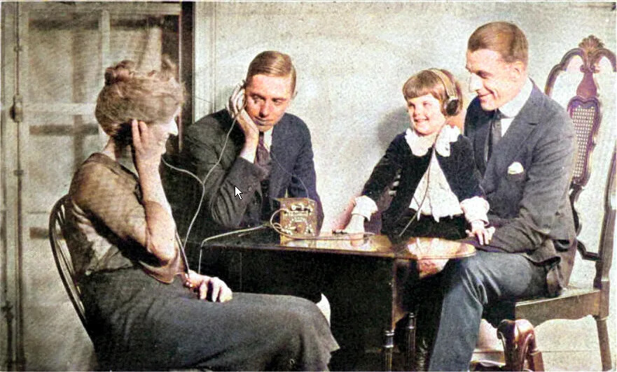 A colored photograph of two men, a woman, and a child sitting around a small brown box on a table. The child, the woman, and one of the men wear headphones attached to the box. Everyone in the photograph is smiling at each other.