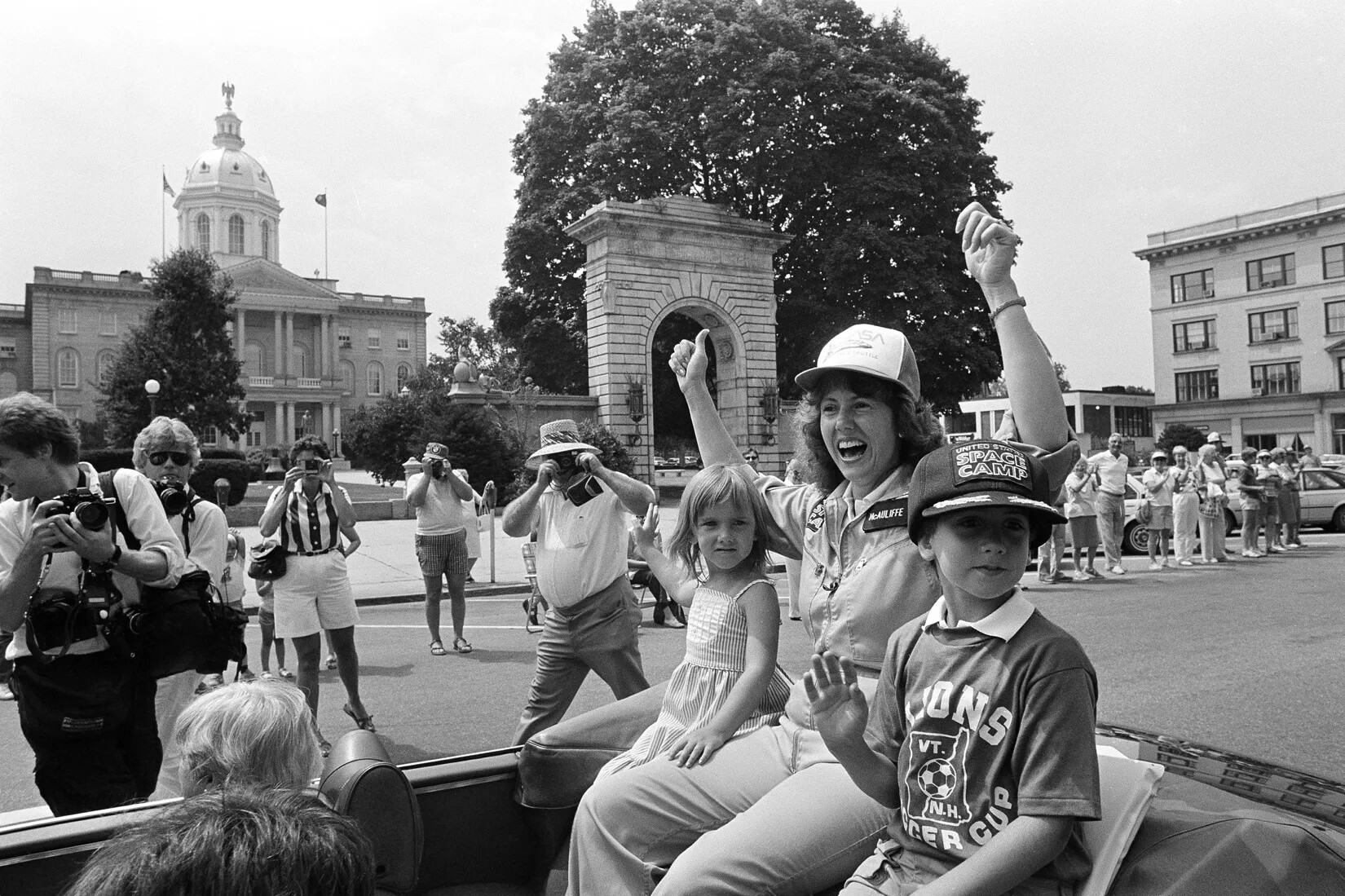 A black and white photograph of a woman and two children riding in an open-top car. The woman has both her arms raised and she is smiling. The two children, a girl to the woman's left and a boy to the woman's right, each wave with one hand. There are crowds of people watching the car pass in the background. In the background of the left-hand side of the photograph is a group of photographers with cameras of varying sizes. Behind this group of people is a large archway and a three-story building with a dome. Two levels of column-lined porches jut out from the center of the building and is crowned by a pointed roof. 