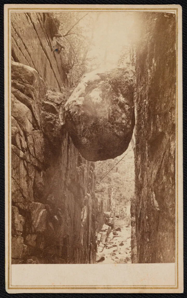 A large boulder is caught between two rock walls in a gorge in a black and white photograph. The walls are steep, and there are some trees in the background. The photograph does not show the ground.