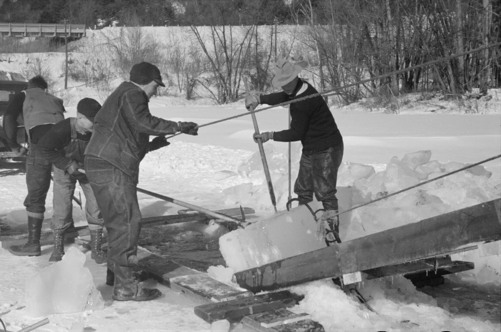 A black and white photograph of four men pulling pieces of ice off a wooden ramp onto a snow-covered surface.